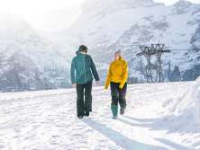 Deux personnes marchent depuis la station amont de First en direction d'Oberjoch sur un sentier de randonnée hivernal enneigé. Le soleil bas crée un contre-jour intense et de longues ombres sur la neige. À l'arrière-plan, on aperçoit des sommets enneigés aux contours saisissants.