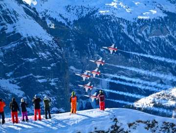 Jets der Patrouille Suisse fliegen in enger Formation über das Lauberhornrennen. Im Vordergrund stehen Zuschauer auf einem verschneiten Hang und verfolgen die Flugshow, während die Flugzeuge ihre Rauchspuren vor der steilen, schneebedeckten Bergflanke ziehen.
