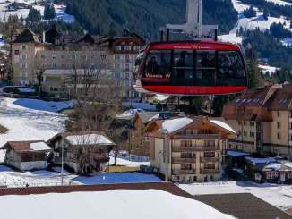 A gondola on the Wengen–Männlichen aerial tramway hovers over the snow-covered village of Wengen. Sports facilities and buildings can be seen below, while the mountain backdrop frames the village in the distance.