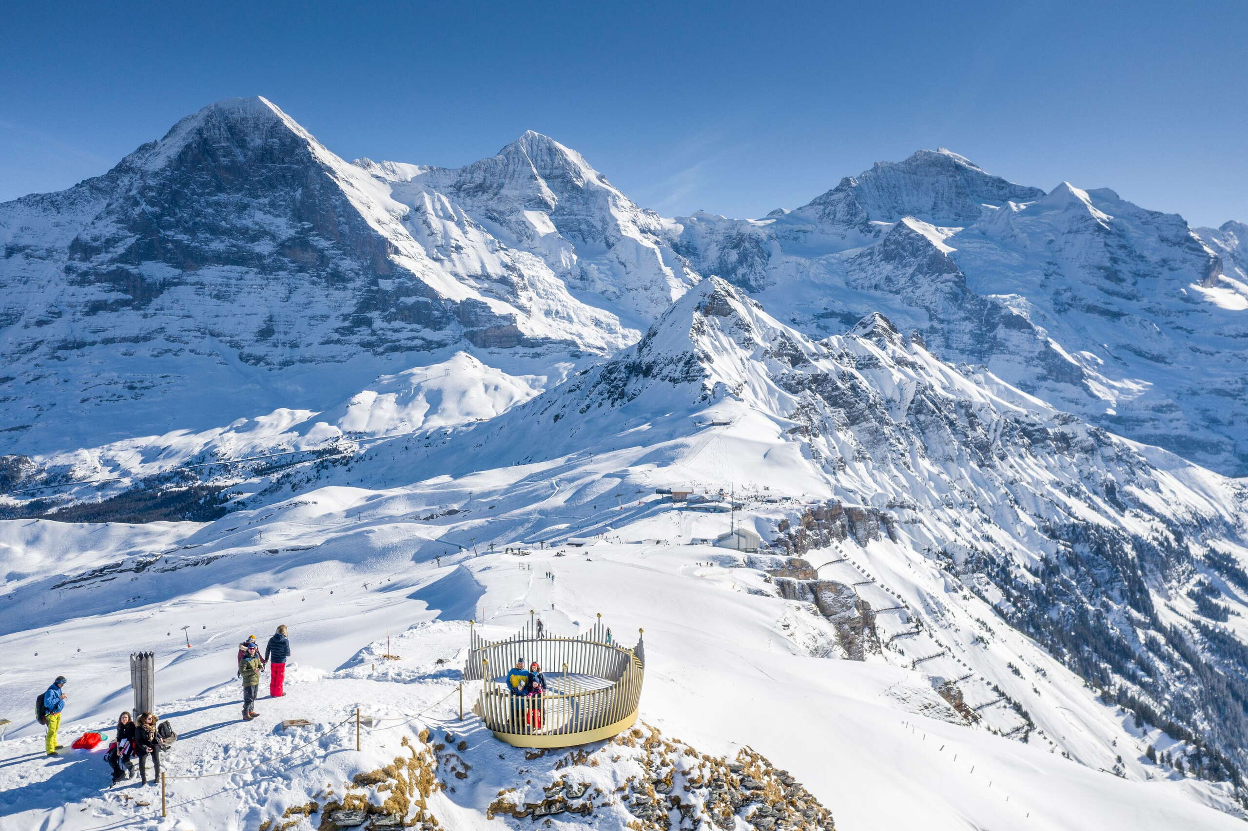 Eine Gruppe von Personen im Tageslicht bei der Station vom Royal Walk. Im Hintergrund die verschneite Bergkette mit Eiger, Mönch und Jungfrau. 