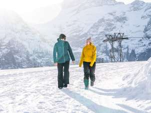 Two people are walking from the First mountain station toward Oberjoch along a snow-covered winter hiking trail. The low sun creates a bright backlight and long shadows in the snow. Striking, snow-capped mountain peaks are visible in the background.
