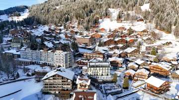 Luftaufnahme des Bergdorfs Wengen mit verschneiten Chalets, Hotels und Strassen. Das Dorf liegt eingebettet am Hang unterhalb eines winterlichen Waldgebiets.