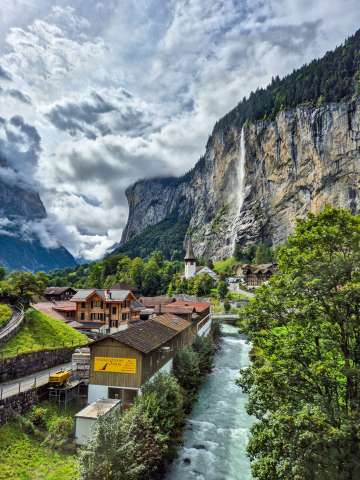 Blick auf das Dorf Lauterbrunnen mit seinen Chalets und der fliessenden Lütschine. Im Hintergrund stürzt der Staubbachfall von der steilen Felswand ins Tal und prägt das Dorfbild.