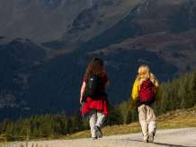 Zwei Wanderinnen gehen auf einem breiten Bergweg durch die offene Landschaft. Vor ihnen erheben sich markante, teils dunkel schattierte Berghänge, die von einzelnen Sonnenflecken und herbstlichen Farbtönen geprägt sind. Links im Schatten fällt der Blick auf Wengen. 