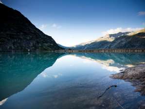 Le lac de Gelmer est niché au cœur du paysage de haute montagne du Grimsel. La surface calme de l'eau reflète les flancs des montagnes environnantes et le ciel limpide. Au premier plan, on aperçoit les rochers des rives, tandis que le barrage délimite le lac à l'arrière-plan.