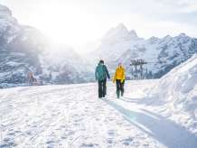 Zwei Personen gehen von der Bergstation First Richtung Oberjoch über einen verschneiten Winterwanderweg. Die tief stehende Sonne sorgt für helles Gegenlicht und lange Schatten im Schnee. Im Hintergrund sind markante, schneebedeckte Berggipfel sichtbar.