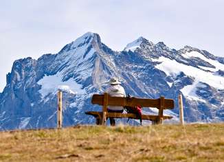 A wooden bench stands in an alpine meadow. Steep, partly snow-capped peaks rise in the background, while the foreground appears open and peaceful.