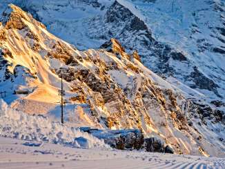 Au premier plan, une piste de ski fraîchement damée, aux traces bien marquées, traverse la haute montagne enneigée. À l'arrière-plan, les flancs rocheux et enneigés de l'Eiger et du Mönch resplendissent dans la lumière chaude du matin.
