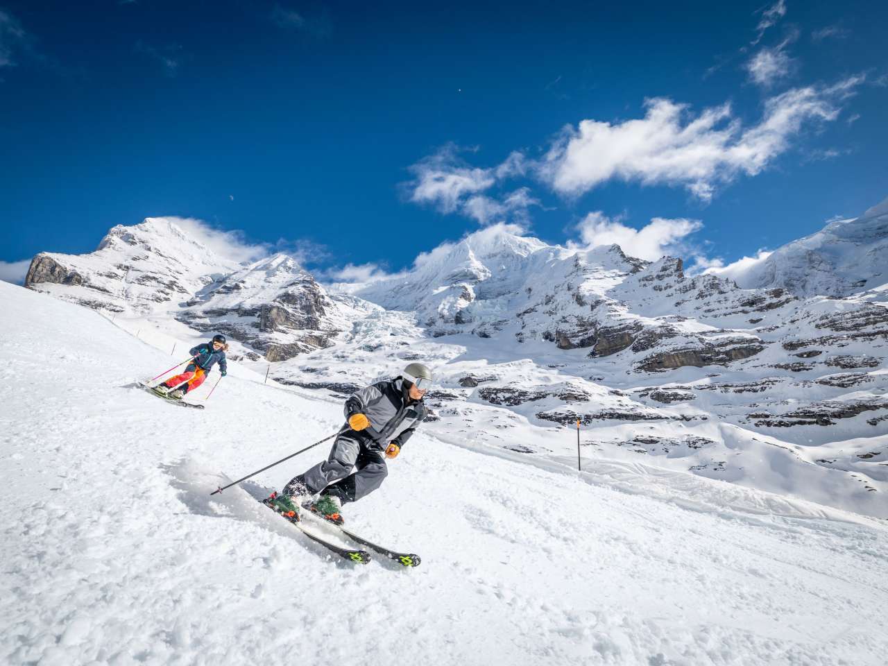 Zwei Skifahrer fahren über eine präparierte Piste. Im Hintergrund ist eine verschneite Bergkulisse zu sehen.