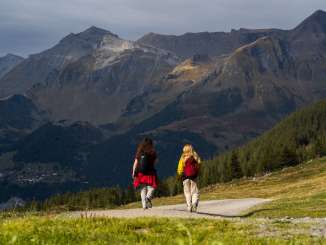 Two hikers are walking along a wide mountain trail through the open countryside. Ahead of them rise striking mountain slopes, some of which are darkly shaded, dotted with patches of sunlight and autumnal hues. To the left, in the shade, Wengen comes into view. 