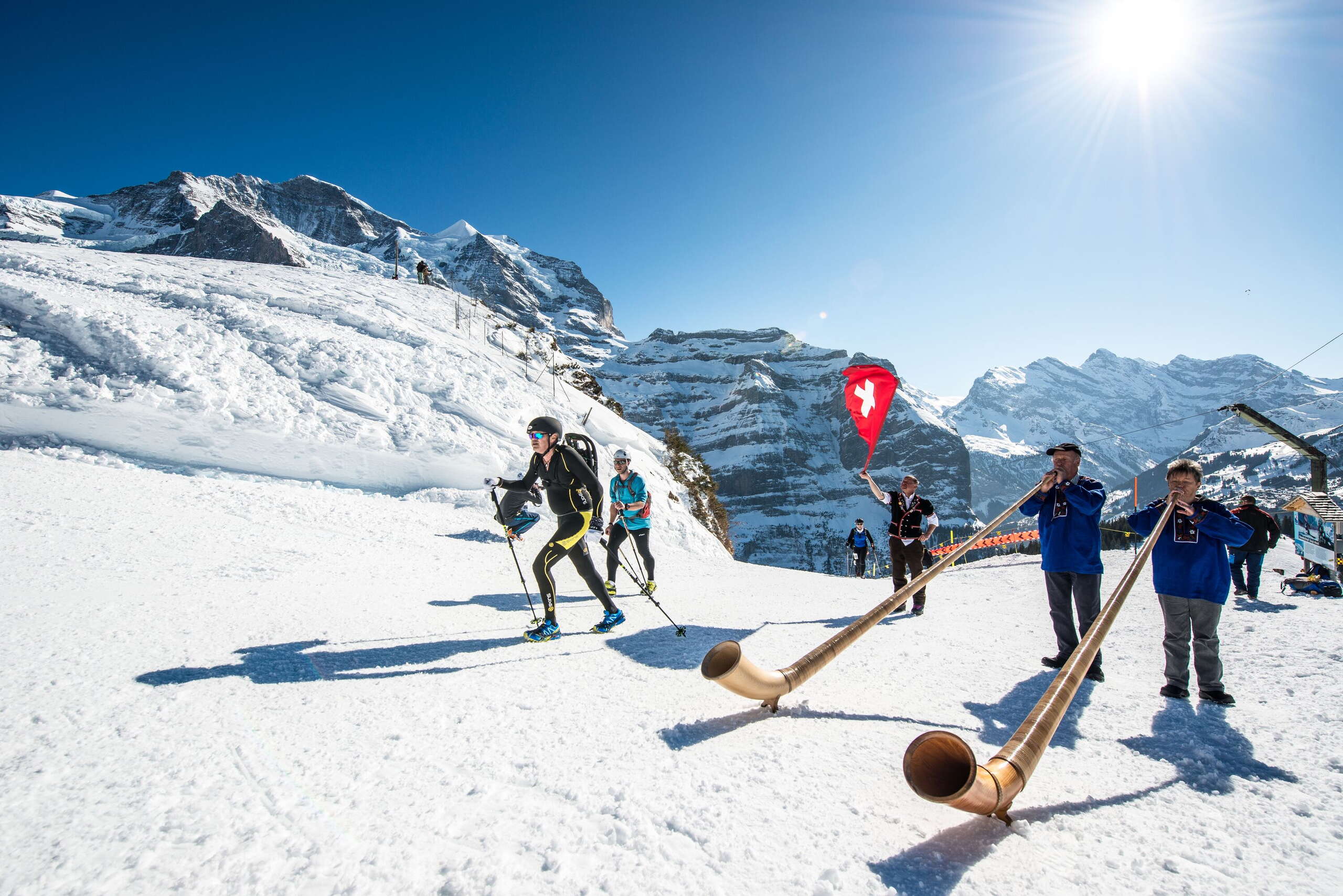 Beim Vertical UP in Wengen sind die Läufer zu Fuss auf dem Schnee unterwegs. Neben der Rennstrecke steht ein Fahnenschwinger und zwei Alphornbläser*innen.