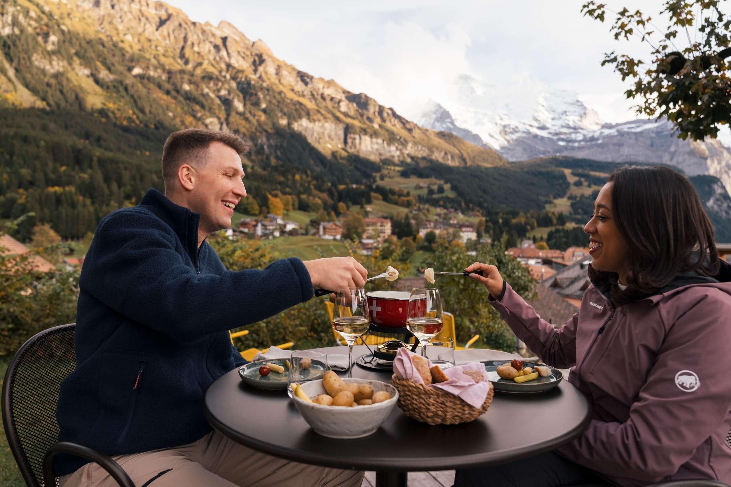 Deux personnes sont assises à une table ronde sur une terrasse et tiennent des fourchettes à fondue trempées dans du fromage fondu. Sur la table se trouvent des assiettes d'accompagnements, des verres à vin, une corbeille à pain et un caquelon à fondue. À l'arrière-plan, on aperçoit des maisons, une forêt et des montagnes.