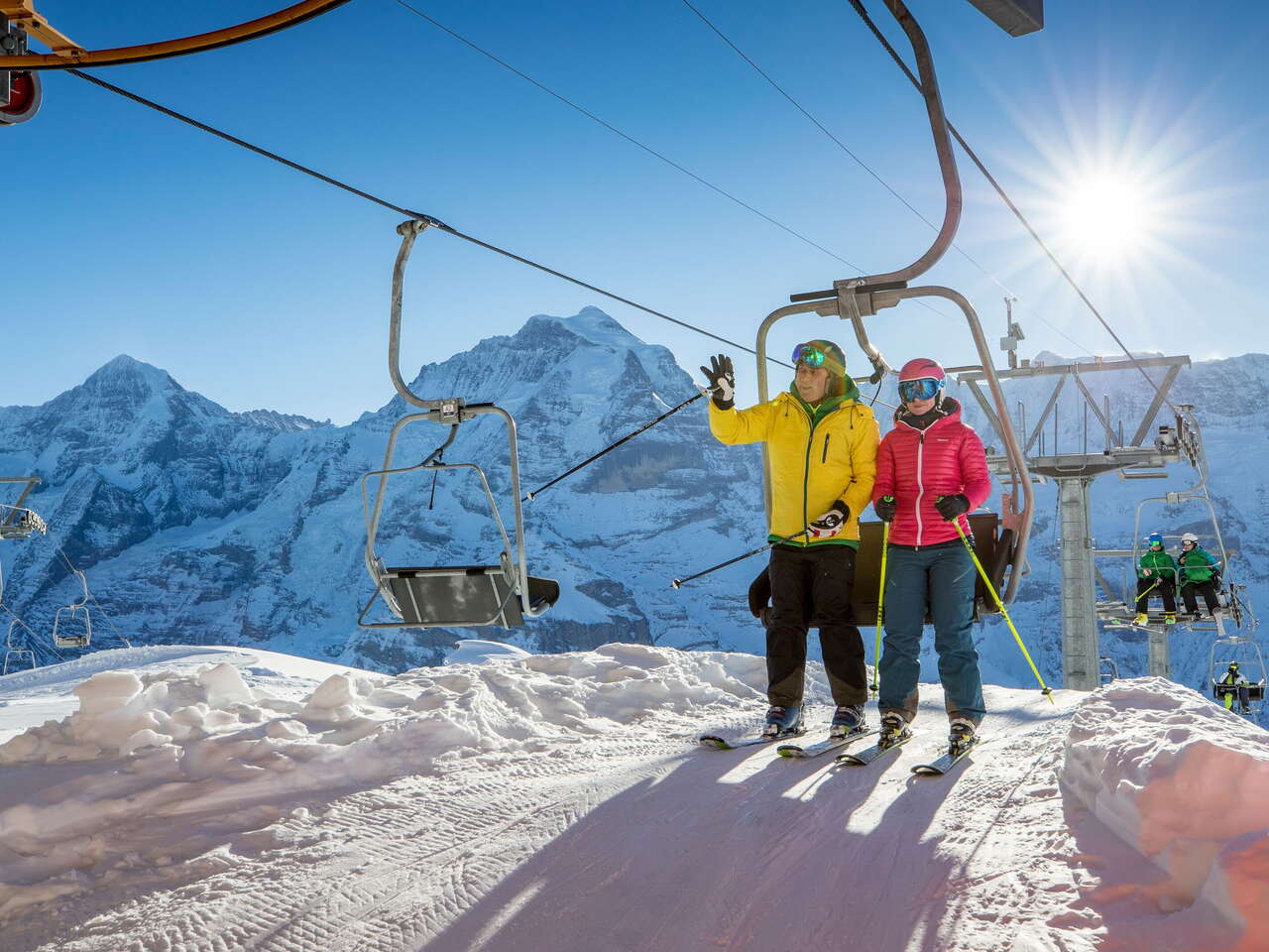 Skifahrer stehen vom Sessellift auf im Skigebiet Mürren-Schilthorn. Das Dreigestirn Eiger, Mönch und Jungfrau ist aus der Ferne zu sehen.